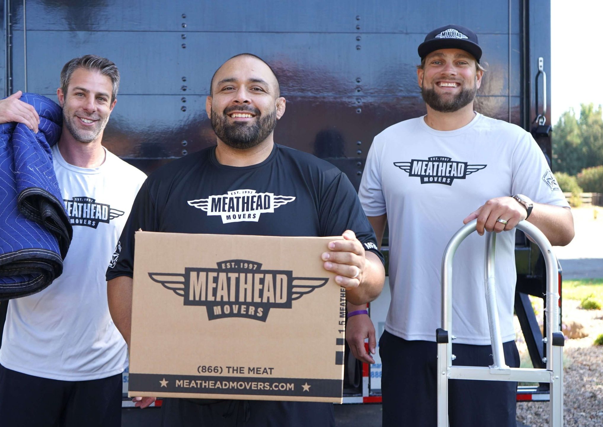 Three Meathead movers holding a moving box and moving blanket beside a truck